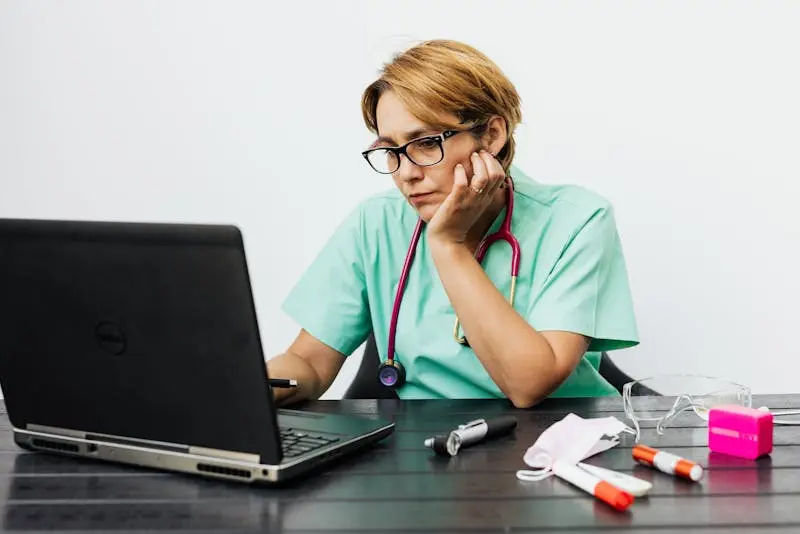 A woman in medical scrubs is at a desk, engaged with her laptop, appearing concentrated.