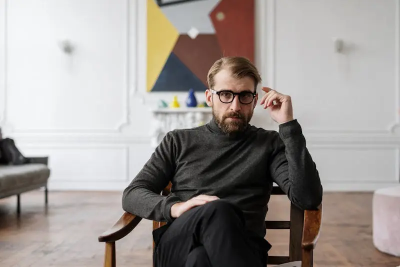 A man wearing glasses sits in a chair, relaxed, in a well-lit room with neutral-colored walls.