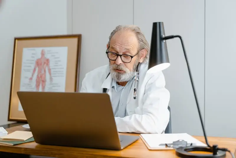 A senior man wearing a white coat is seated at a desk, focused on his laptop.