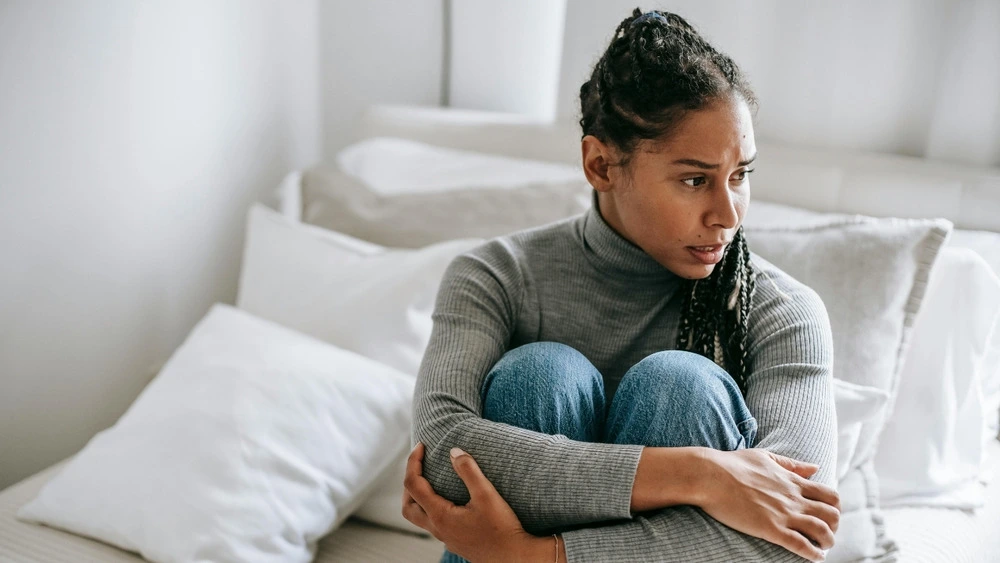 A woman sitting on a bed, hands resting on her knees, looking contemplative.